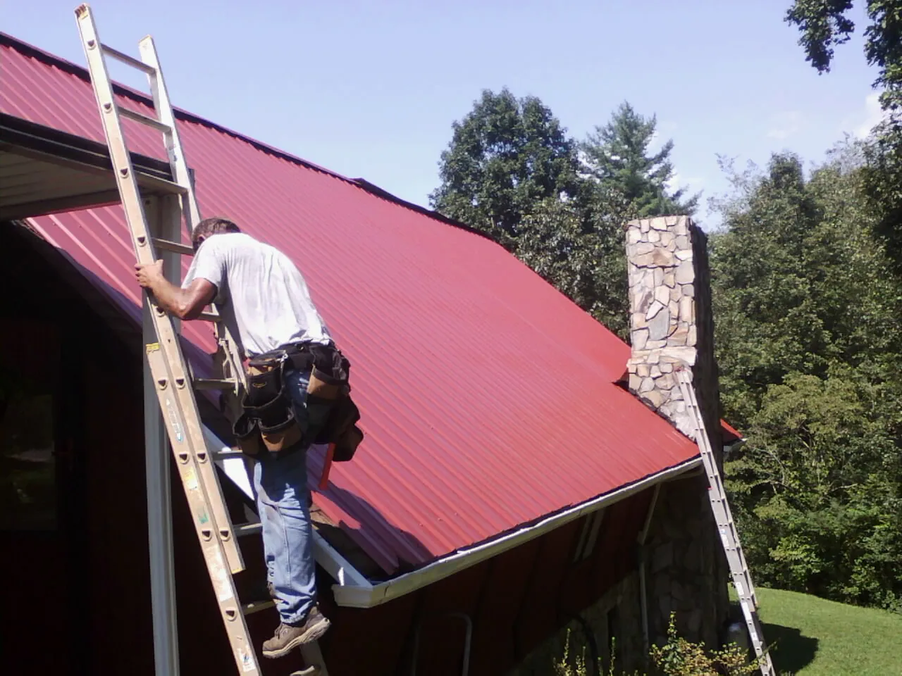 Roofer on a ladder beside a red metal roof and stone chimney — Soffit & Fascia Repair by Roofing Brothers in Randallstown